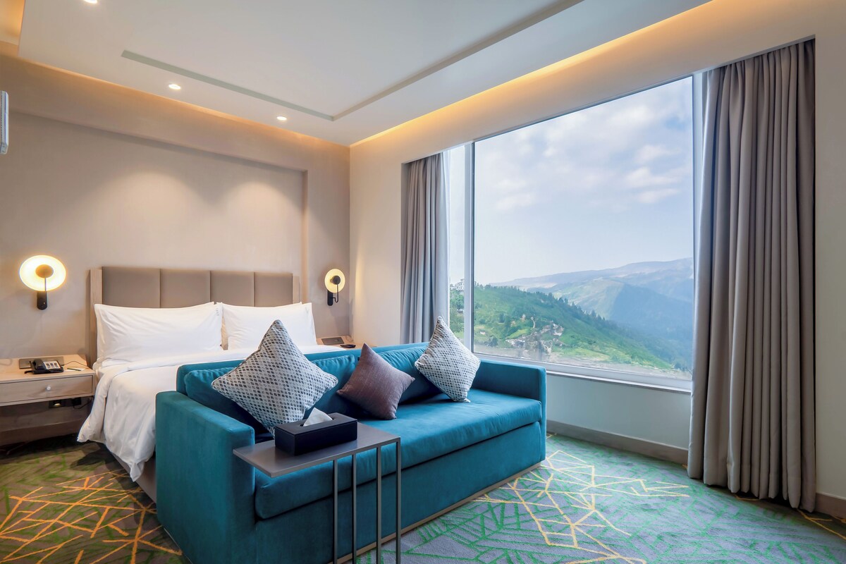 A hotel room with a bed, a blue sofa, a nightstand, and a large window with a view of mountains.