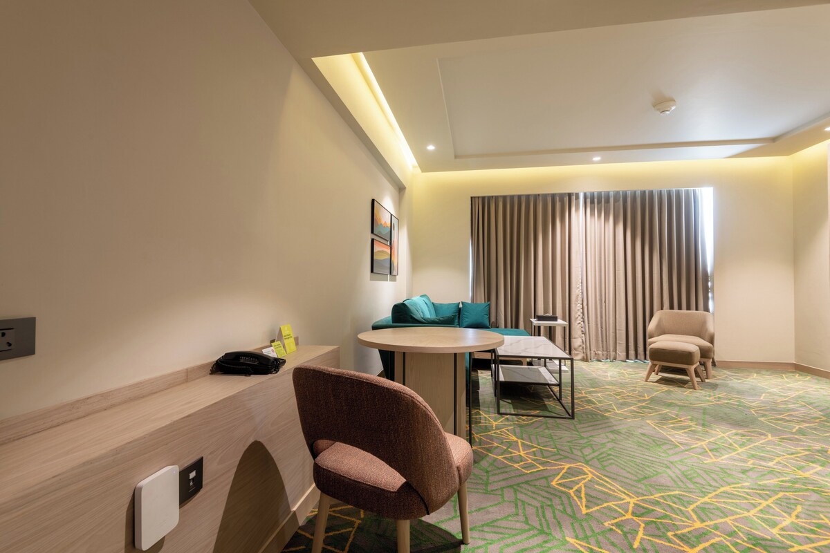 Habitación de hotel con una mesa redonda, un sofá, una silla, un escritorio con teléfono y una ventana con cortinas.