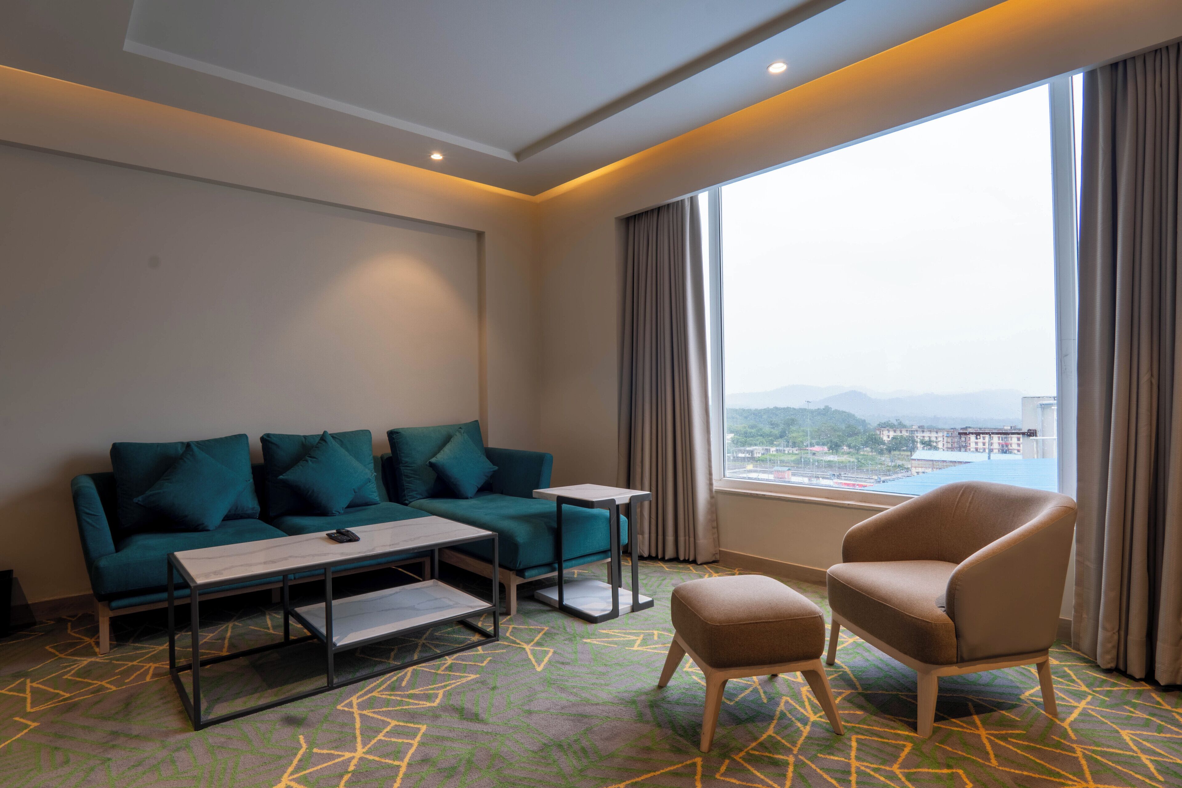 Habitación de hotel con sofá verde, sillón color beige, una mesita y vistas a un paisaje urbano a través de amplios ventanales.