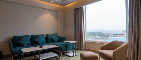 Habitación de hotel con sofá verde, sillón color beige, una mesita y vistas a un paisaje urbano a través de amplios ventanales.