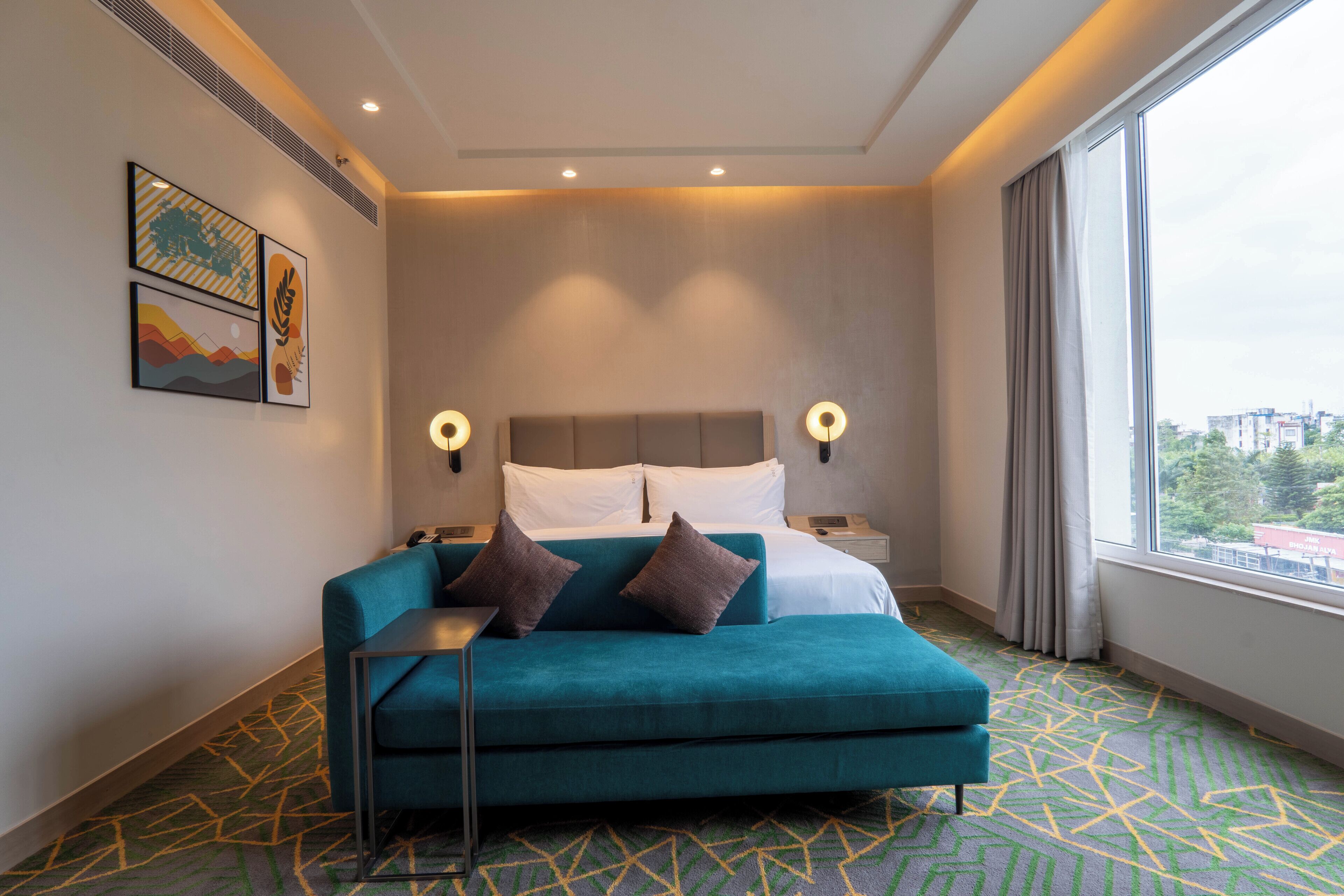 A modern hotel room with a teal sofa, a bed with white linens, two wall-mounted lamps, and a large window with a city view.