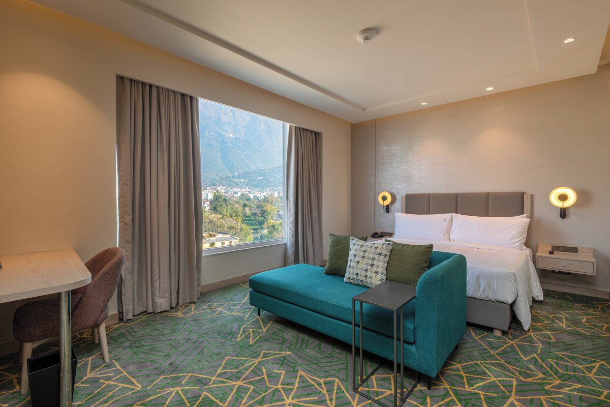 Habitación de hotel con cama, sofá, escritorio y vistas a un paisaje montañoso.