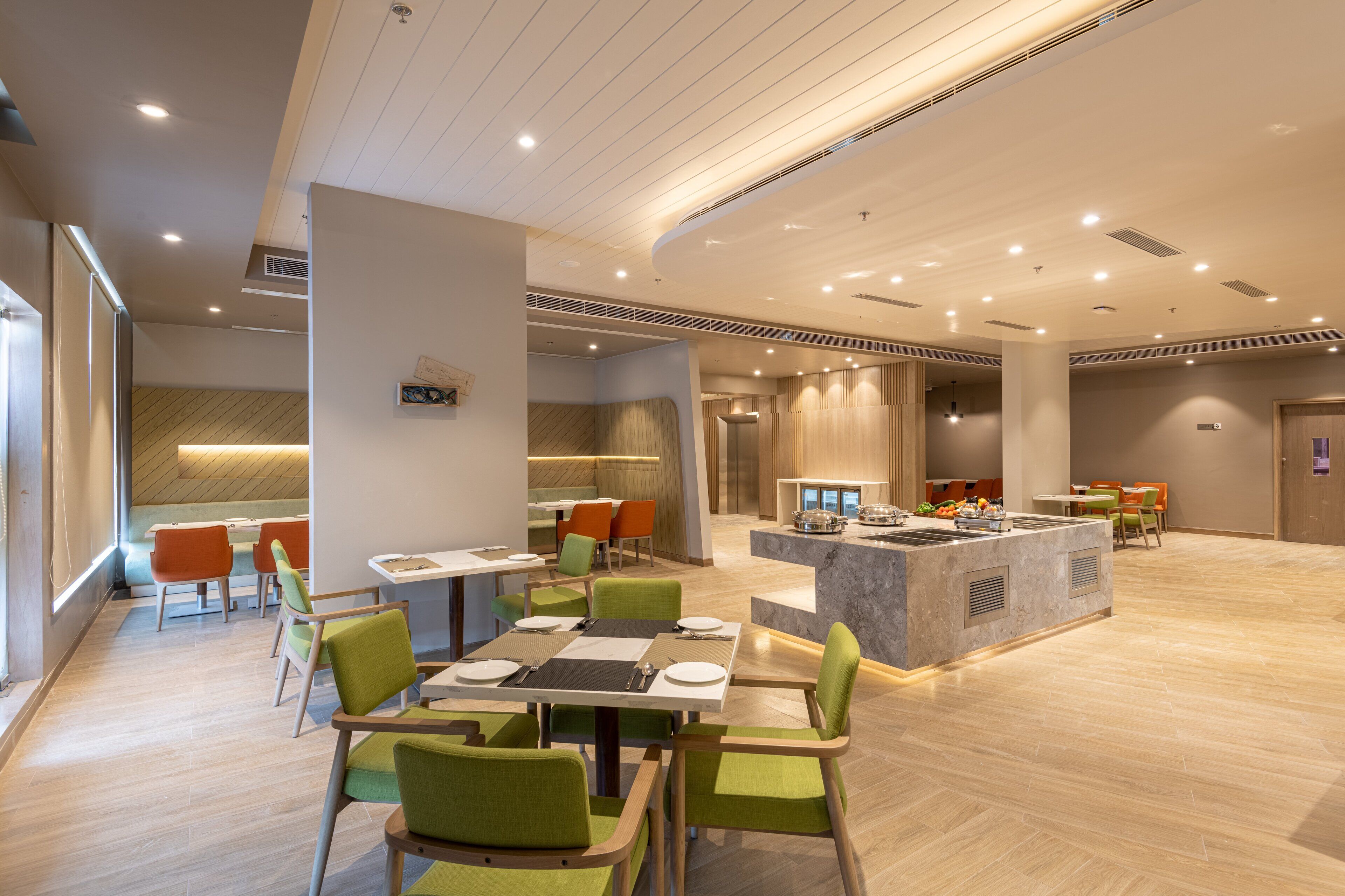 A modern hotel lobby with a central dining area, a reception desk, and a seating area with green chairs.