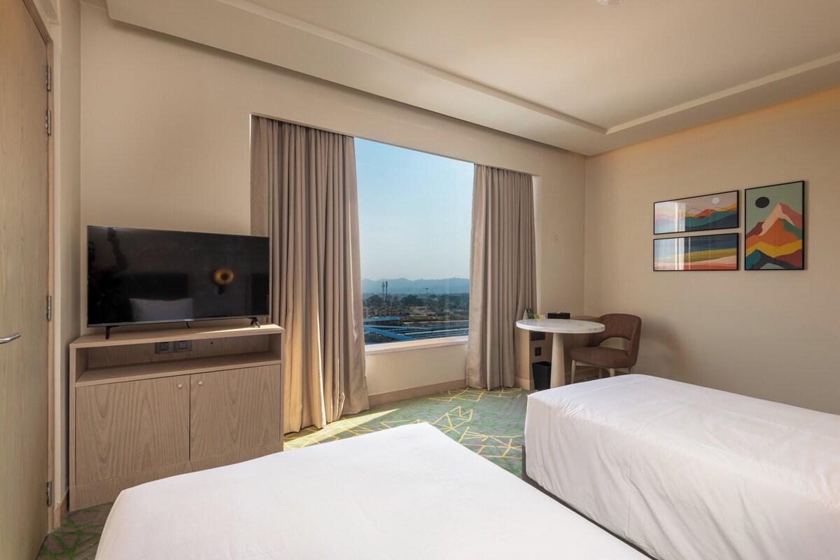 Habitación de hotel con dos camas, un televisor, una mesita y ventana con vistas.