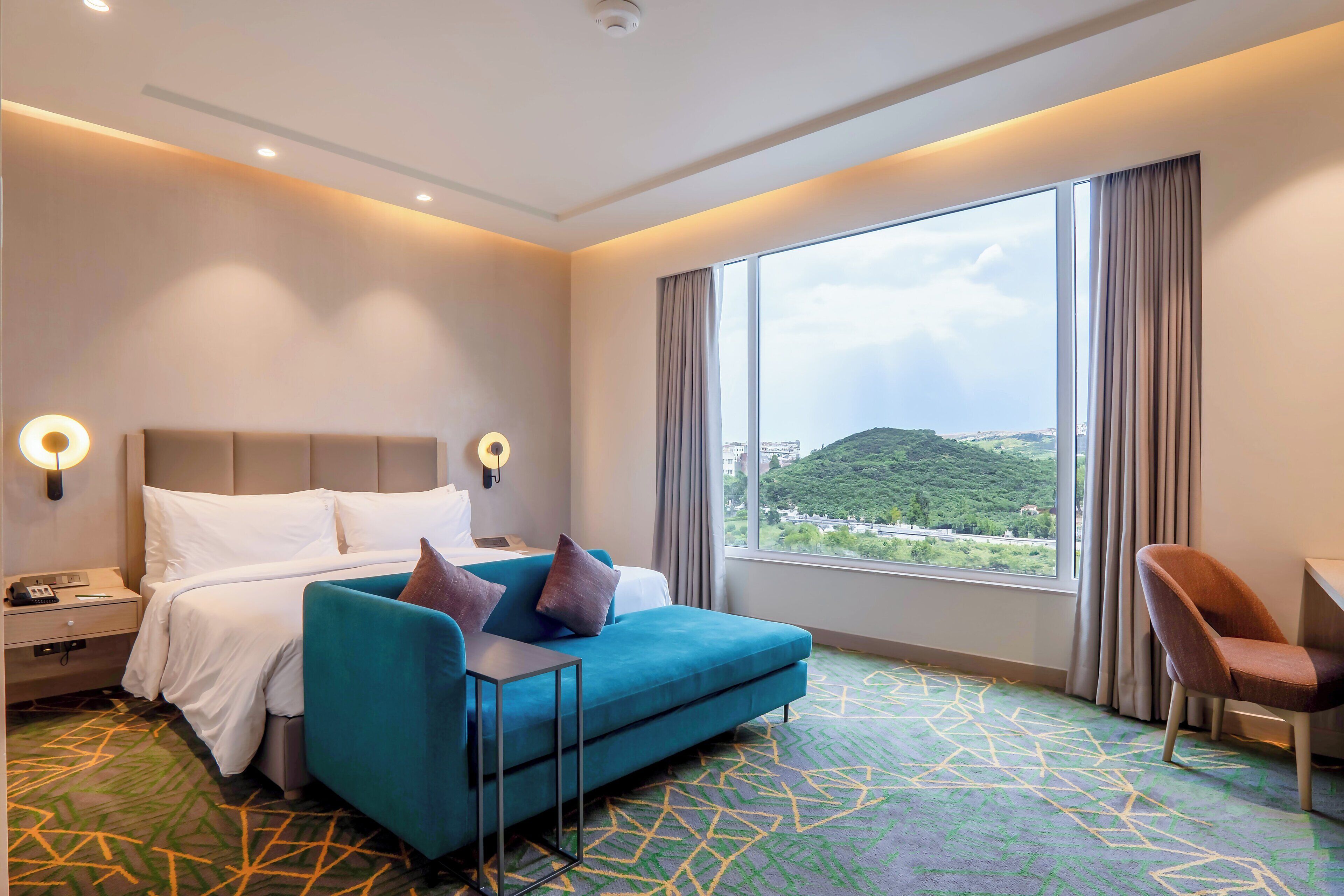 A hotel room with a bed, a blue sofa, a desk, and a large window with a view.