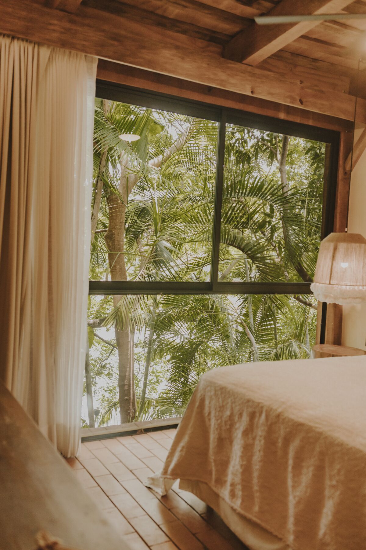 Una habitación con un ventanal grande, vigas de madera, una cama con cubrecama color beige y una lámpara sobre una mesita de noche de madera.