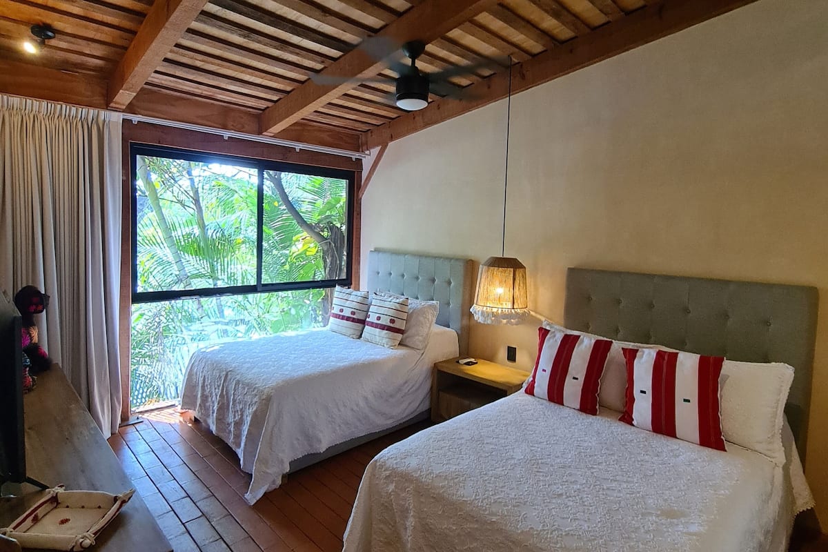 Habitación con dos camas, piso de madera, ventilador de techo, ventana con vista a la vegetación y un escritorio con silla.
