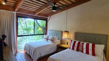 A room with two beds, wooden flooring, a ceiling fan, a window with a view of greenery, and a desk with a chair.