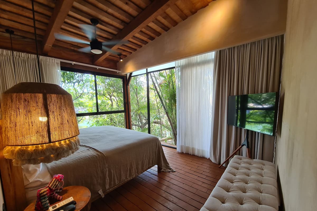 Un dormitorio con cama, televisor, ventilador de techo y ventanas amplias.