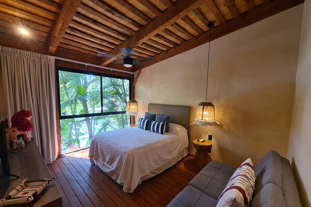 Un dormitorio con techo de madera, una cama con ropa blanca, un sofá gris y una mesita de noche con una lámpara.