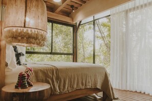A bedroom with a bed, a hanging lamp, a small table, and a view of trees through large windows.