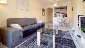 Una sala de estar moderna con un sofá gris, una mesa de centro de vidrio y un comedor con sillas blancas y una mesita.