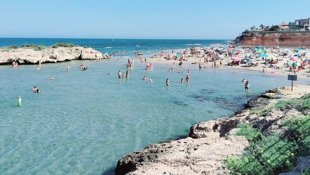 Una playa concurrida, agua cristalina, costa rocosa y sombrillas.
