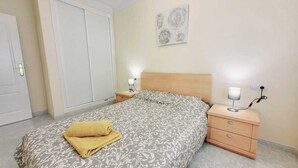 Un dormitorio con una cama, dos mesitas de noche, una mesita auxiliar, un aire acondicionado instalado en la pared y una puerta.