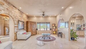 A spacious living area with a stone wall, a white sofa, a round coffee table, and a view of the kitchen through an archway. - Hacienda Foothills Mansion (Tucson)