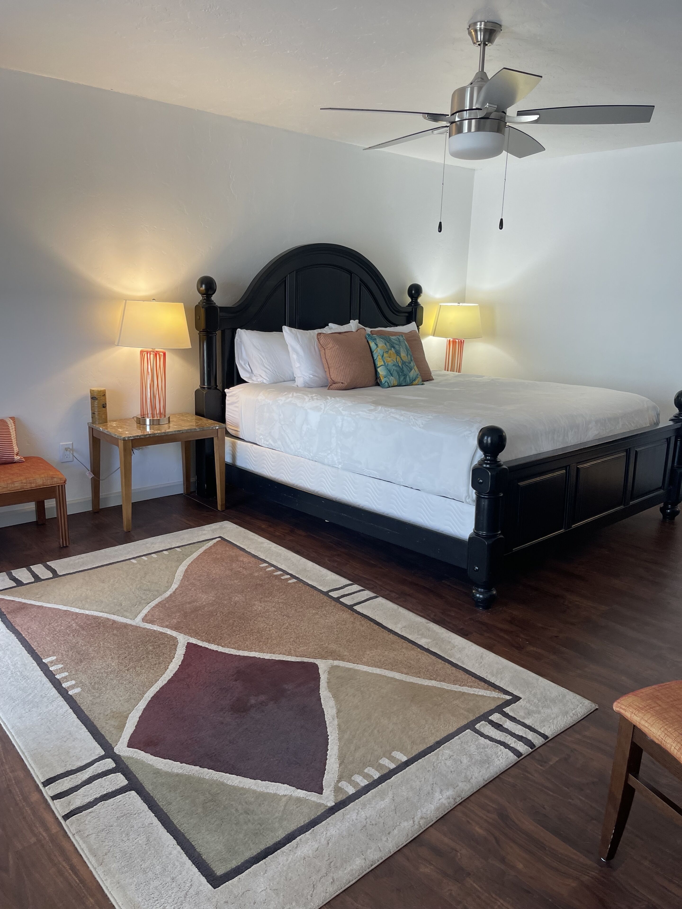 A neatly arranged bedroom with a bed, two bedside lamps, a ceiling fan, and a patterned rug.