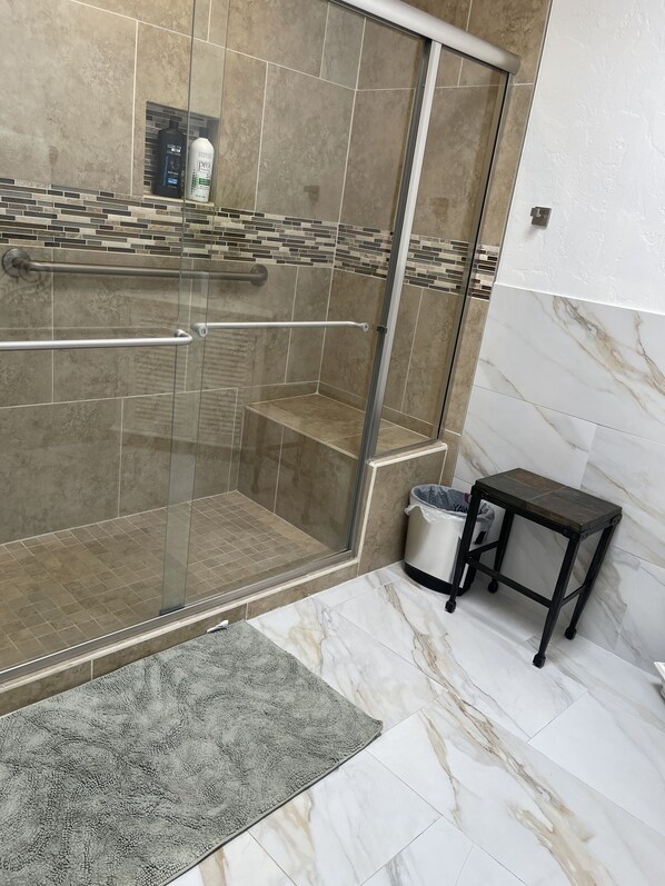 A shower area with a glass door, a tiled floor, a small table, and a trash bin.