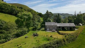 Une maison en pierre avec une cour clôturée, entourée de collines verdoyantes et d’arbres.