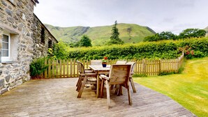 Un bâtiment en pierre doté d’une terrasse en bois, d’une table avec des chaises et offrant une vue sur des collines verdoyantes.