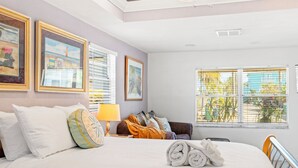 A hotel room with a large bed, a sofa, a window with blinds, and framed artwork on the wall.