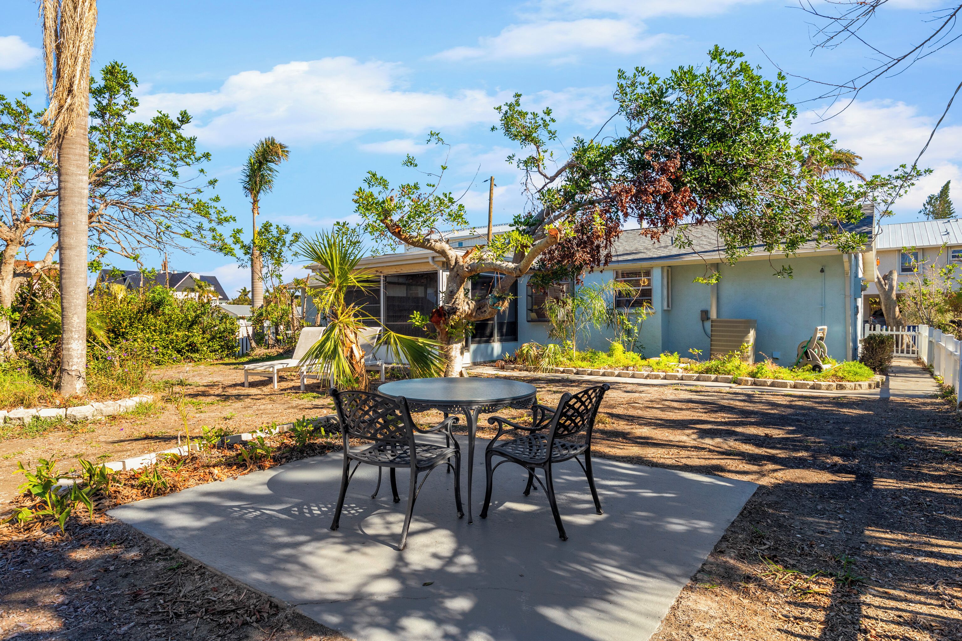 A patio with a round table and four chairs, surrounded by palm trees and a house with a light blue exterior.