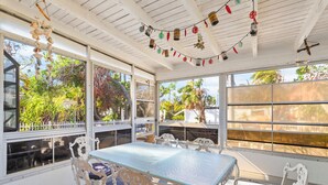 A covered patio with a table and chairs, large windows, and decorative hanging items.