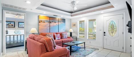 A spacious living room with a large sofa, a coffee table, and a ceiling fan. There is a door with a stained glass window and a painting on the wall.