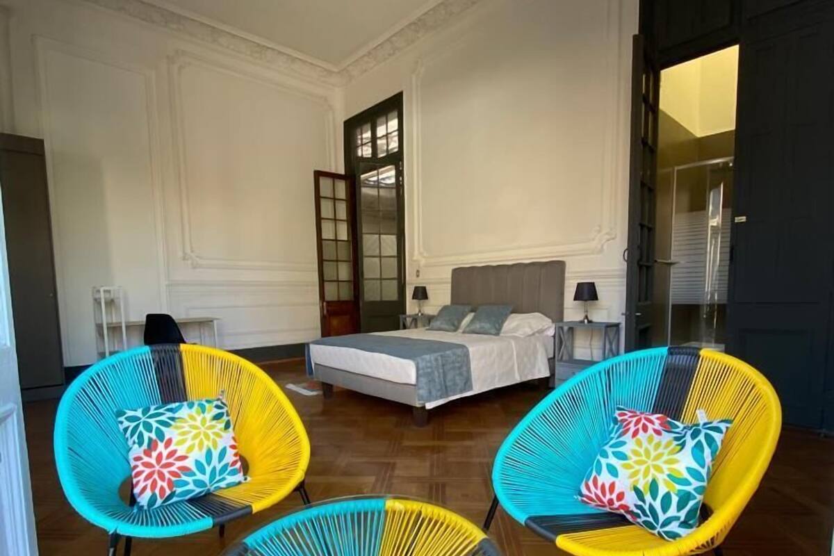 Una habitación con una cama, dos sillas coloridas y una puerta que da a otra estancia.