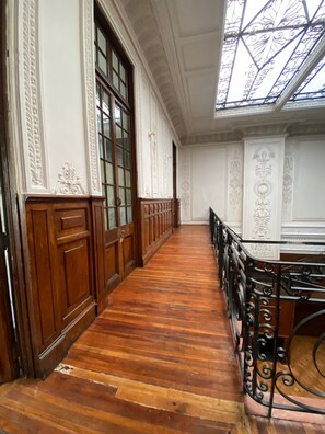 Un couloir aux murs lambrissés et doté d’un plafond en verre, qui mène à une pièce ornée d’une rambarde en métal noir.