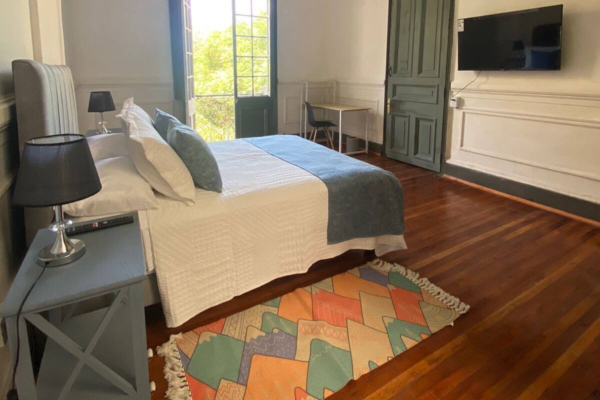 Un dormitorio con cama, mesita de noche, lámpara, televisor y ventana con vistas a zonas verdes.