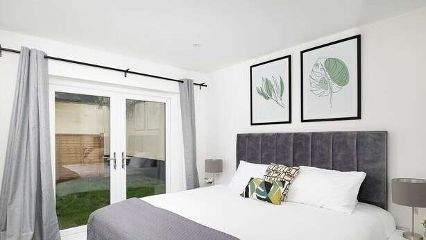 A bedroom with a large bed, grey curtains, a grey headboard, and two framed botanical prints on the wall.