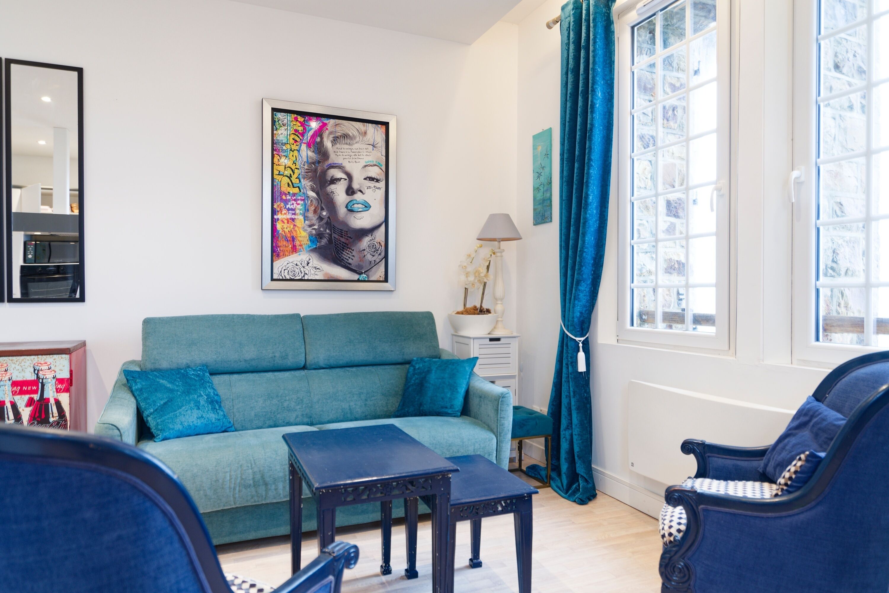 A living room with a teal sofa, a coffee table, and a blue armchair. There is a large framed artwork on the wall and a window with blue curtains.