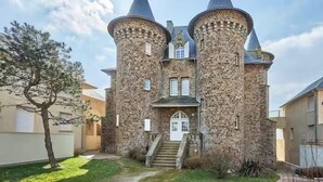 Un bâtiment qui ressemble à un château, avec deux tours imposantes, une entrée centrale avec un escalier et un petit jardin.