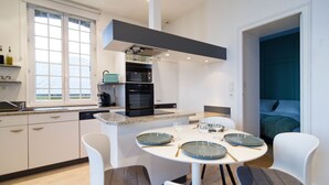 Une cuisine moderne avec des meubles blancs, un îlot central doté de sièges et un coin repas avec des chaises blanches et des assiettes bleues.