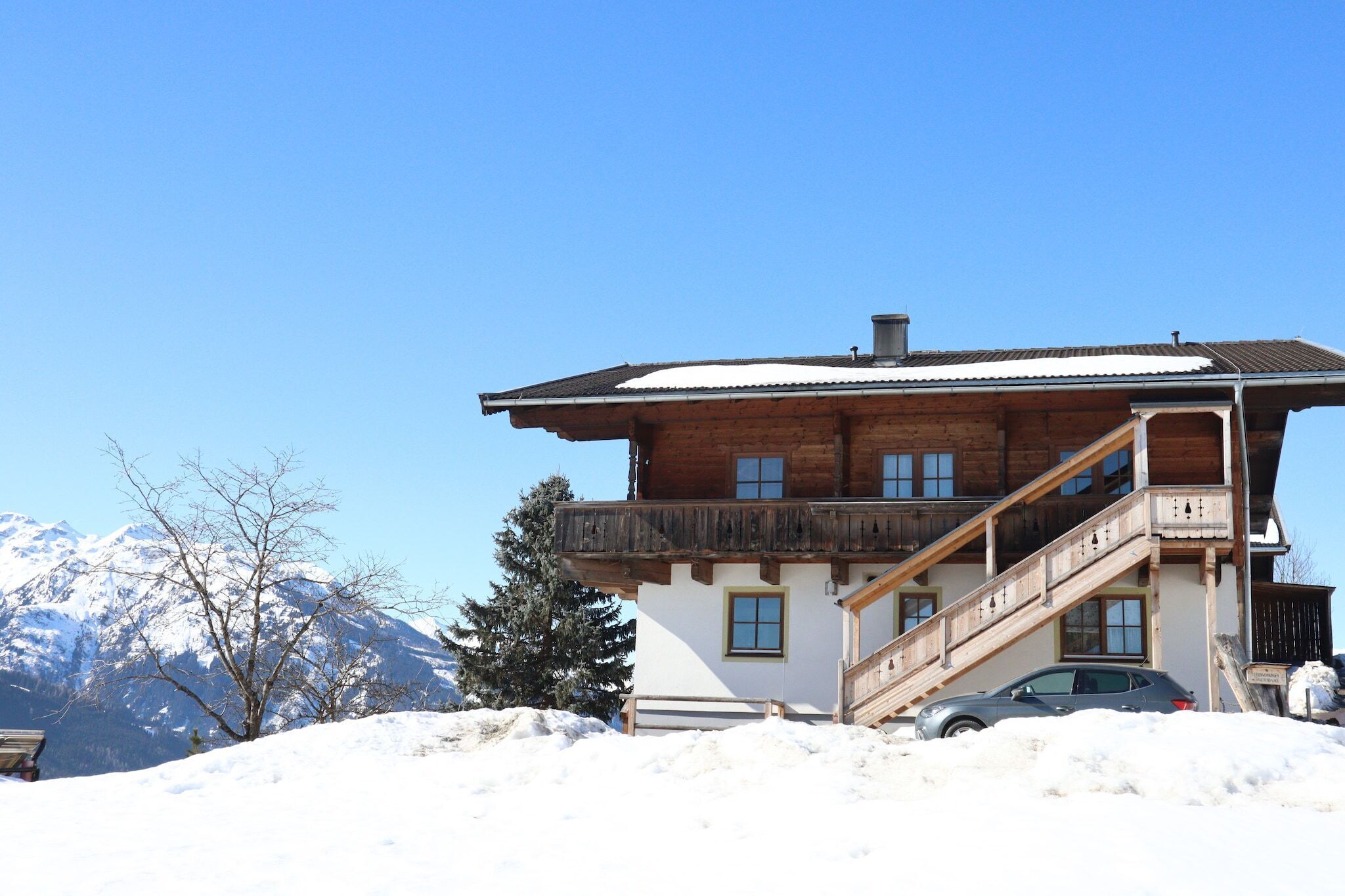 A two-story wooden chalet with a balcony, a car parked in front, and snow-covered mountains in the background.