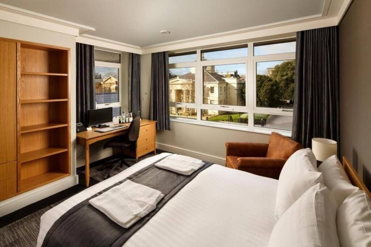 Une chambre d’hôtel avec un lit, un bureau, une chaise, une télévision et une grande fenêtre donnant sur des maisons.