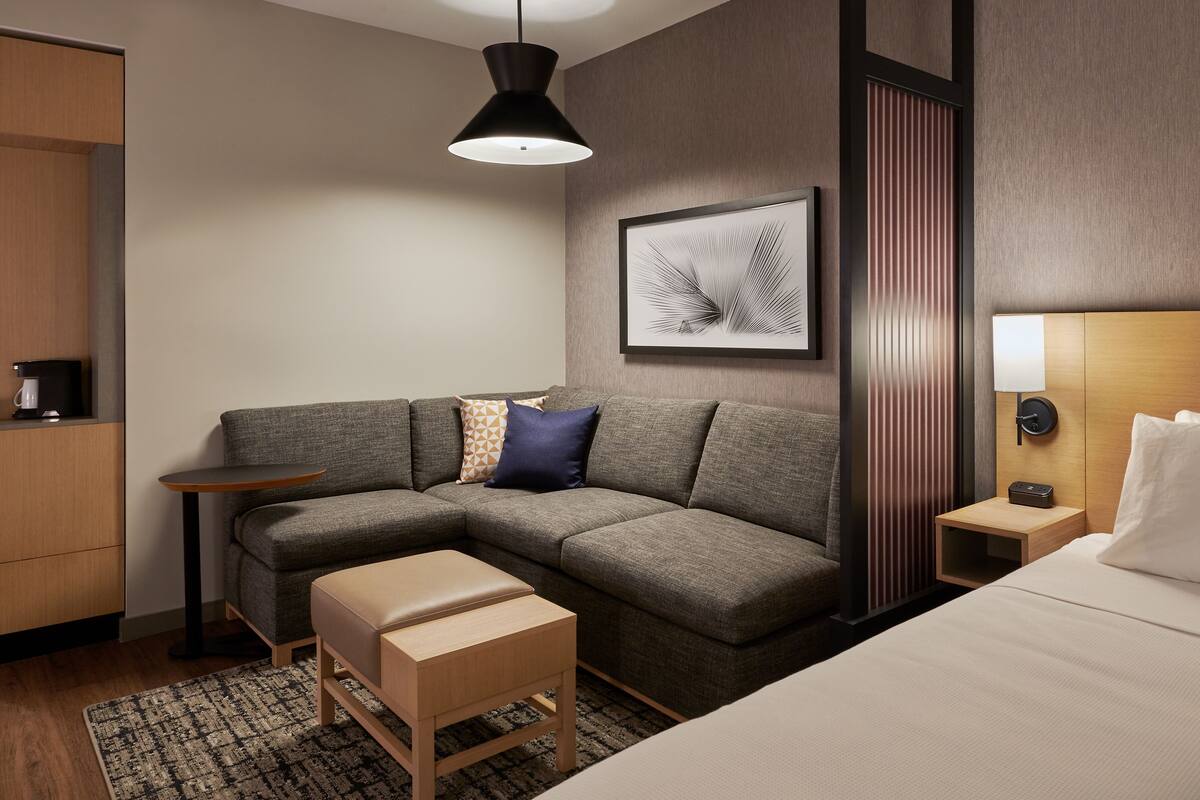 A modern hotel room with a gray sectional sofa, a wooden bench, a small table, a bedside table with a lamp, and a framed artwork on the wall.