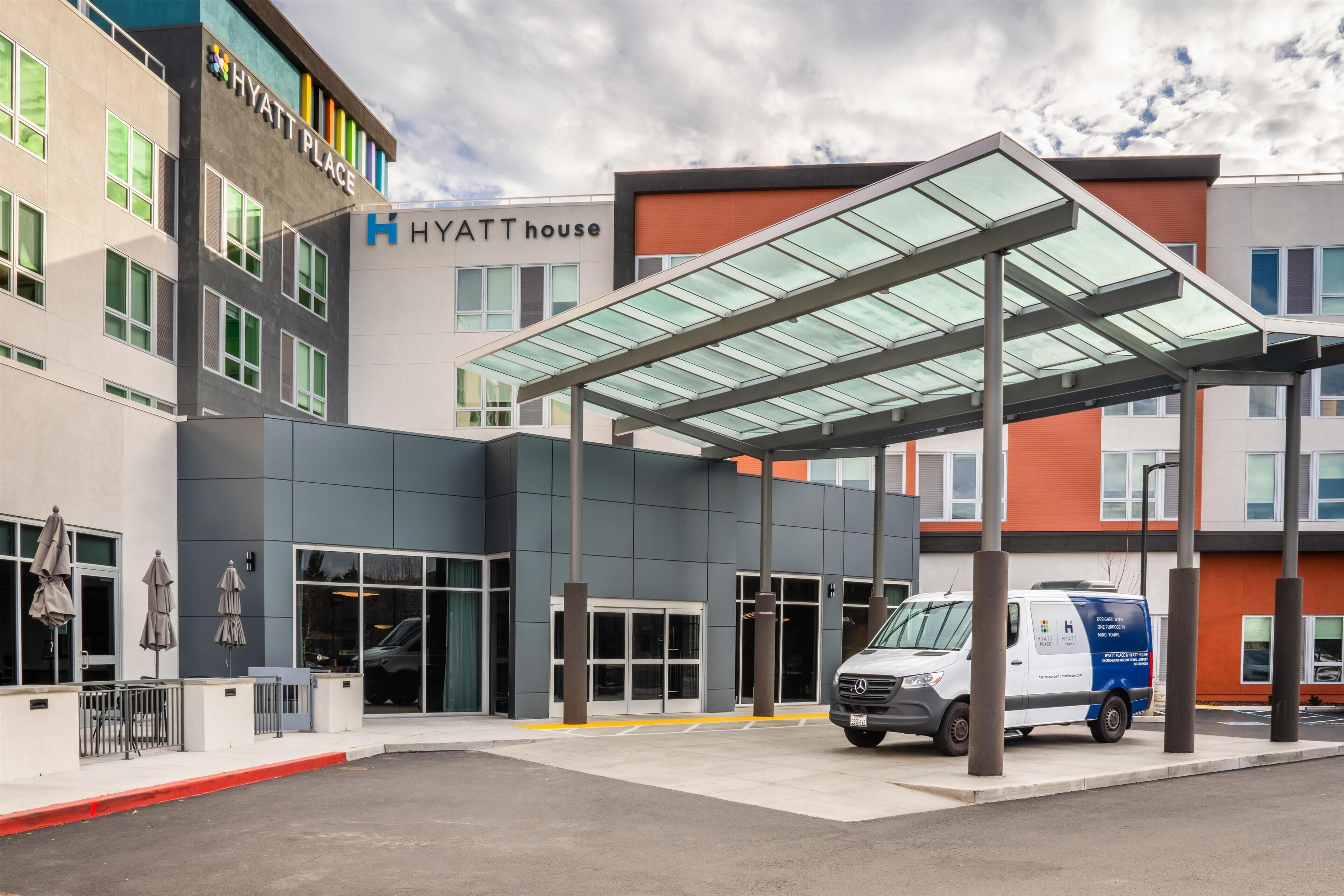 A modern hotel entrance with a covered waiting area, a van, and a clear sky.