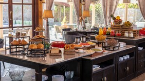 A buffet with various food items and beverages, including pastries, fruits, and drinks, set up in a well-lit room with large windows and curtains. - Brij Bageecha Jaipur - Private Villas with Heated Plunge Pool (Jamwa Ramgarh)