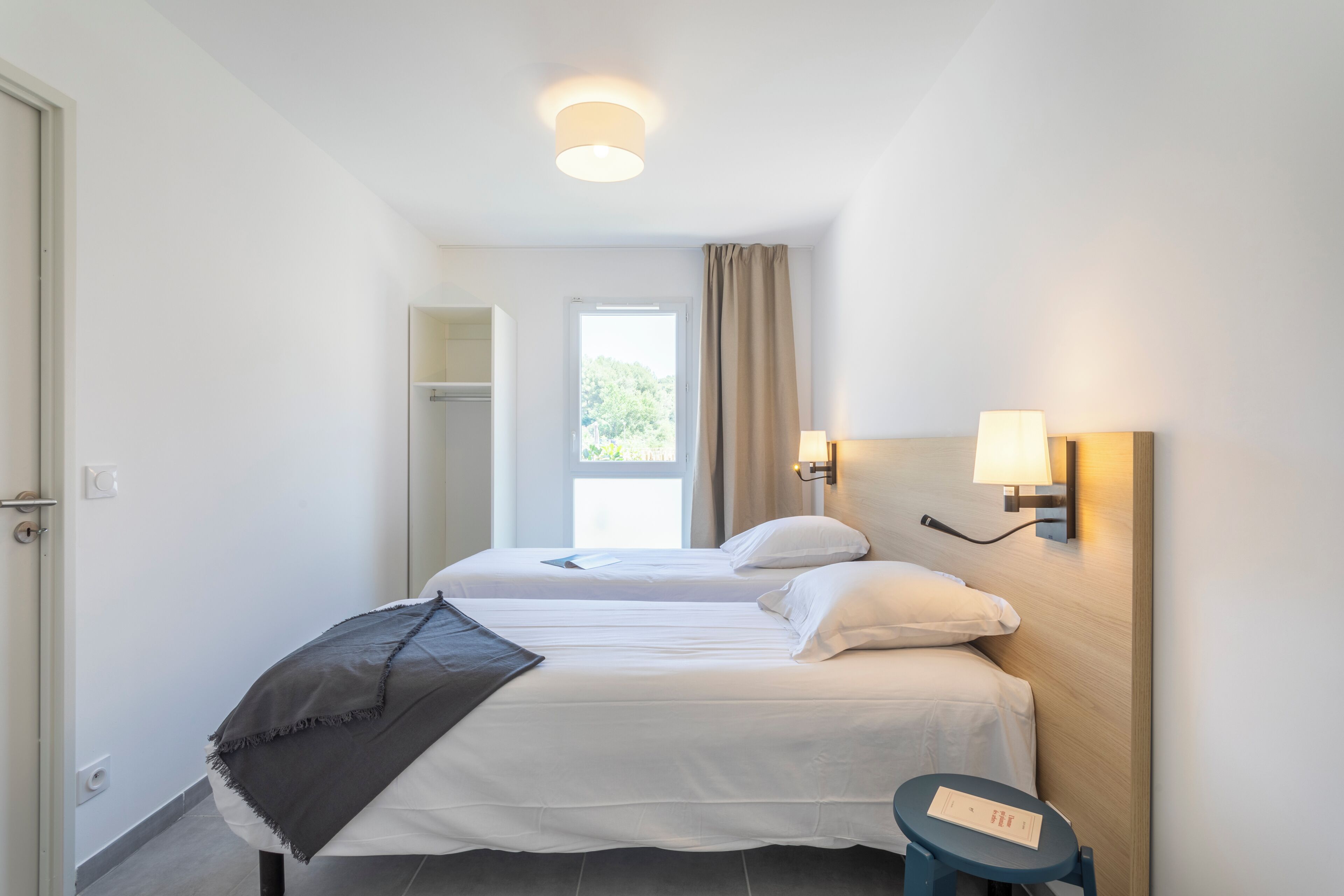 Une chambre d’hôtel avec deux lits, une petite table, une fenêtre avec des rideaux et une lampe fixée au mur.