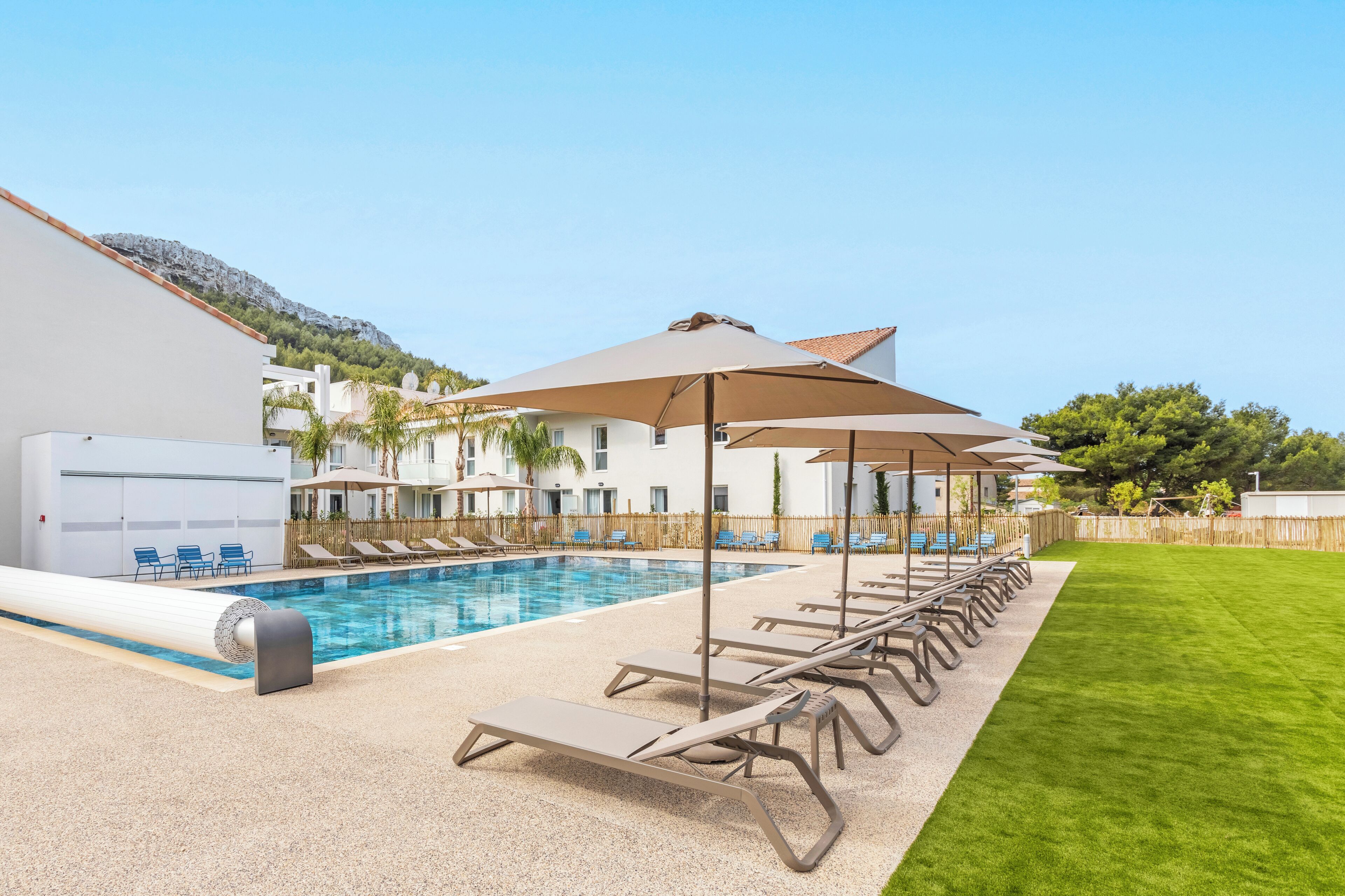 A swimming pool with lounge chairs and umbrellas, surrounded by a well-maintained lawn and a building with a mountain backdrop.