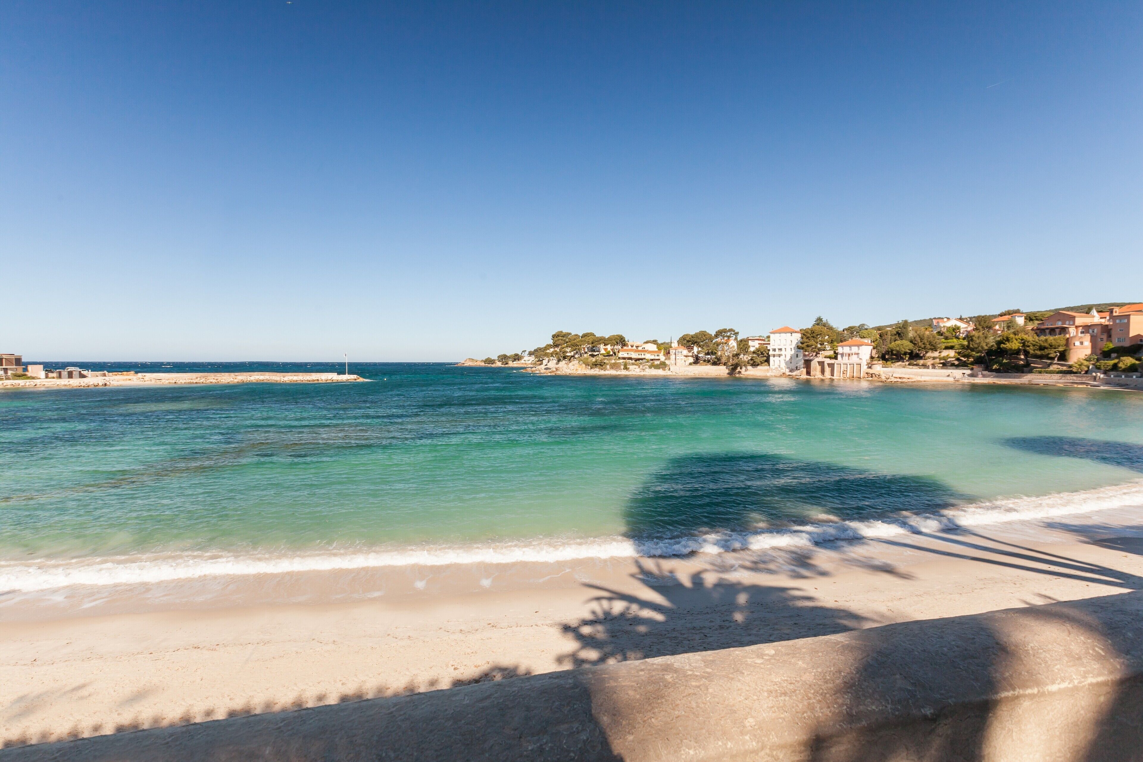 A beach with clear turquoise water, a sandy shore, and buildings in the background.