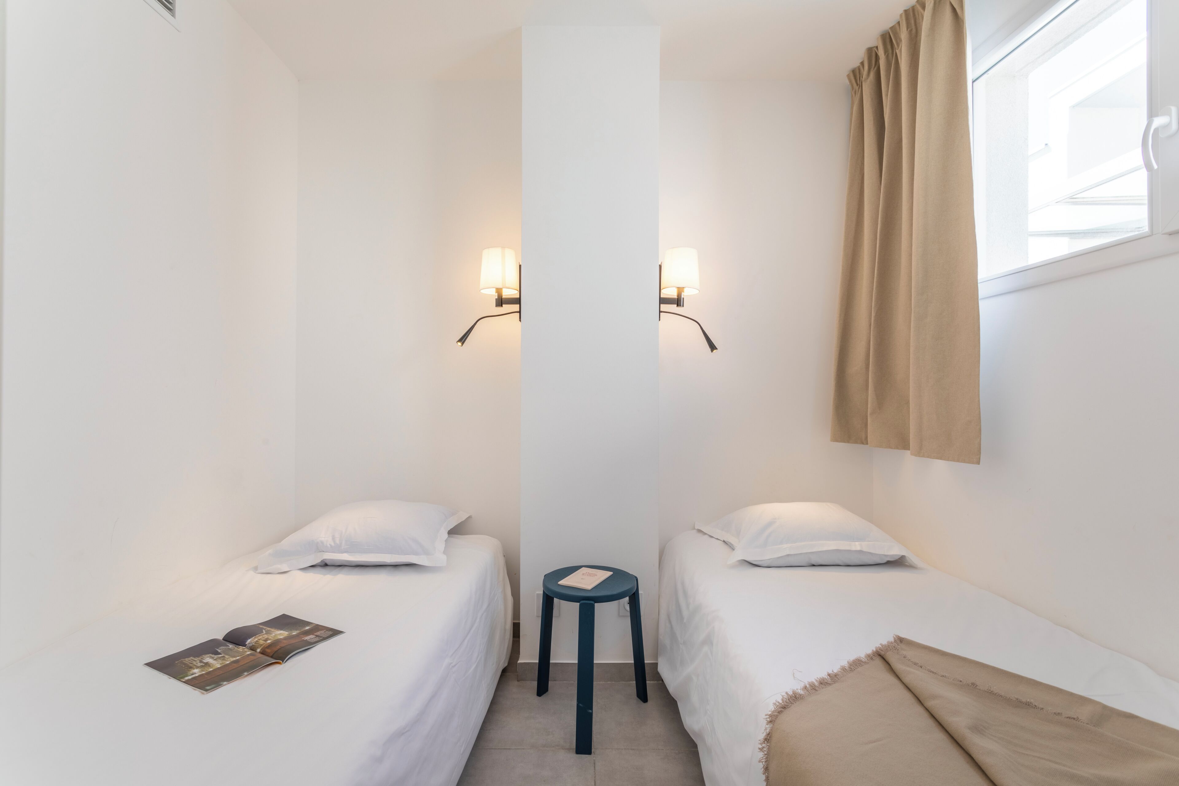 Een kleine, schone kamer met twee eenpersoonsbedden, een klein krukje en twee wandlampen.