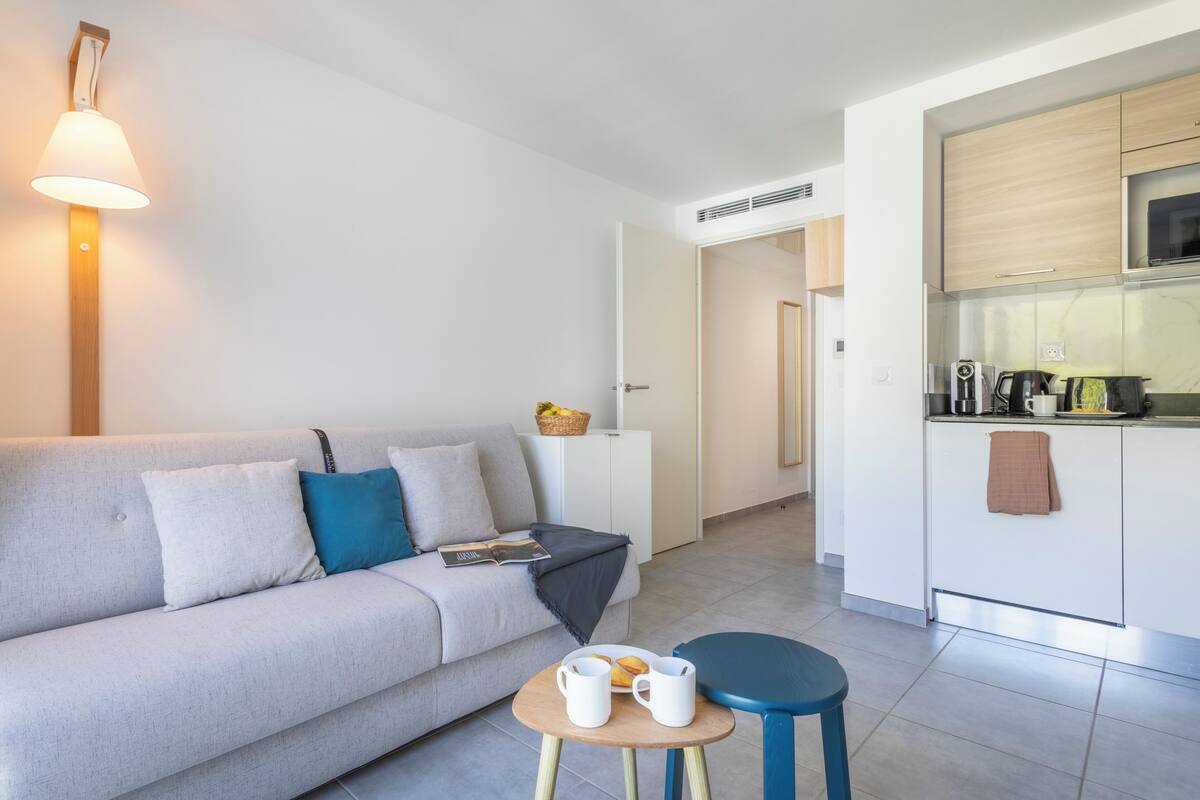 A modern living room with a sofa, a small table with cups and a plate, a kitchen area with cabinets and a microwave, and a hallway leading to another room.