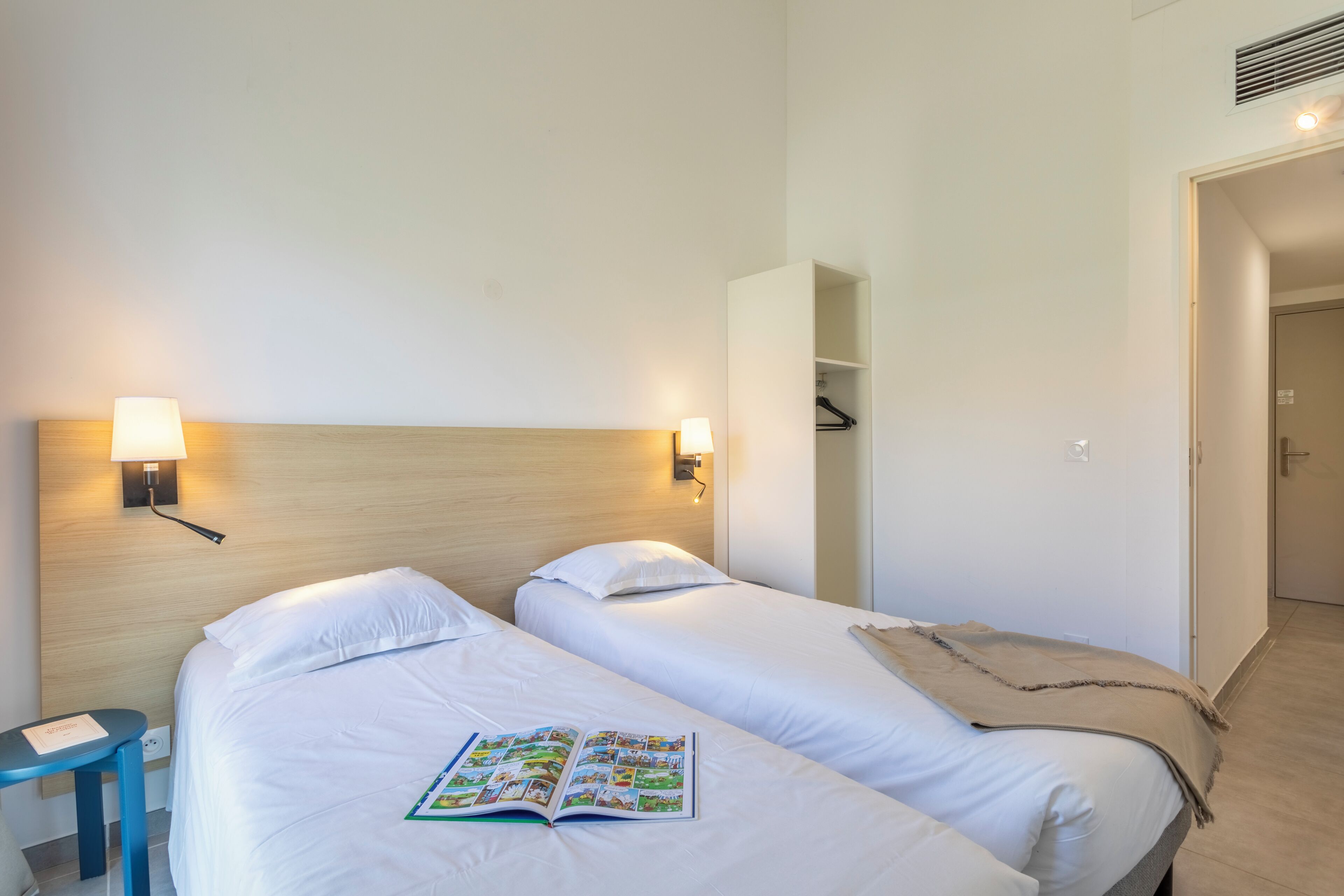 Hotellihuone, jossa on kaksi sänkyä, yöpöytä, lamppu ja ovi, joka johtaa toiseen huoneeseen.