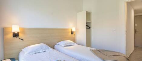 Hotellihuone, jossa on kaksi sänkyä, yöpöytä, lamppu ja ovi, joka johtaa toiseen huoneeseen.