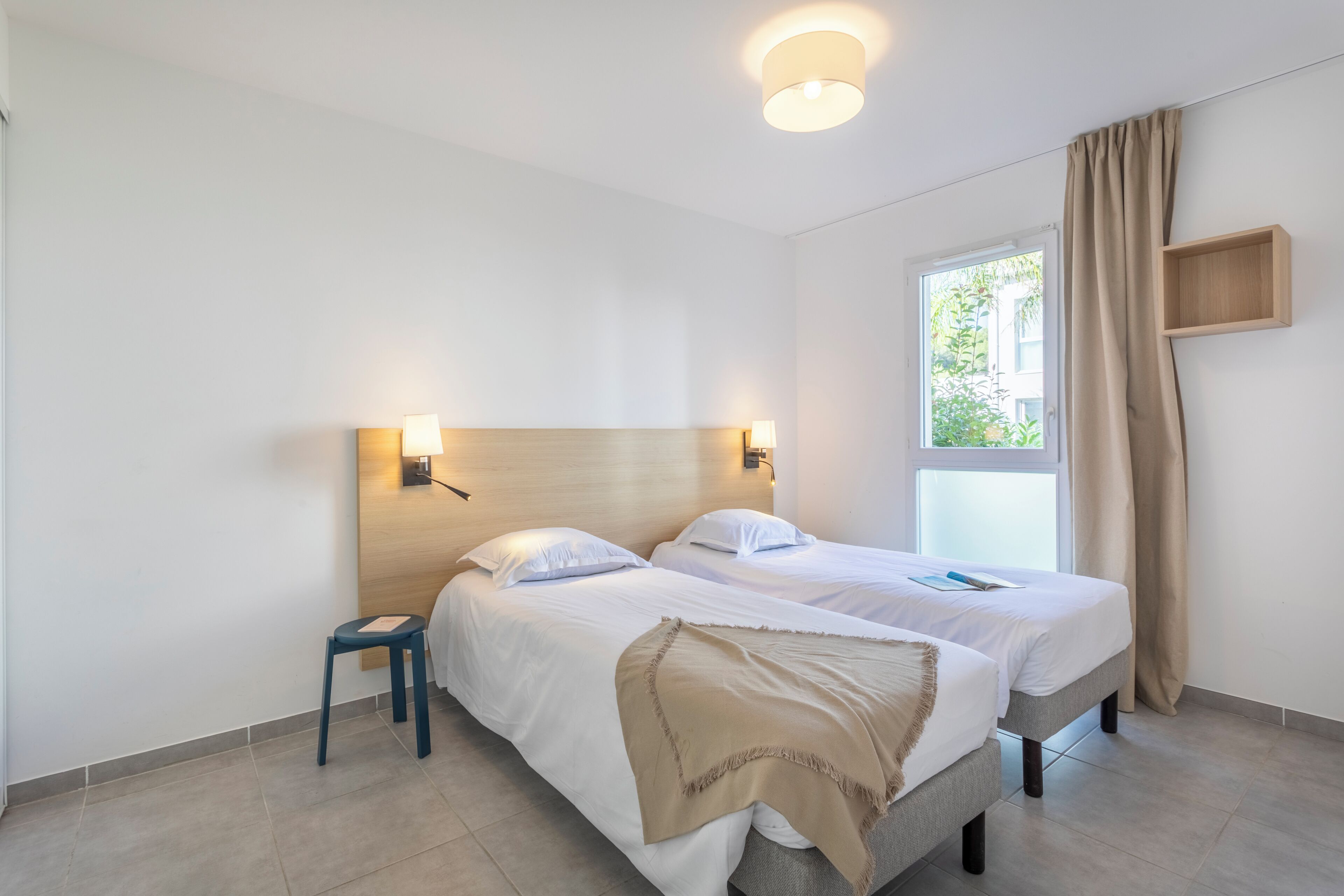 A hotel room with two beds, a small stool, a window with curtains, and a wall-mounted shelf.