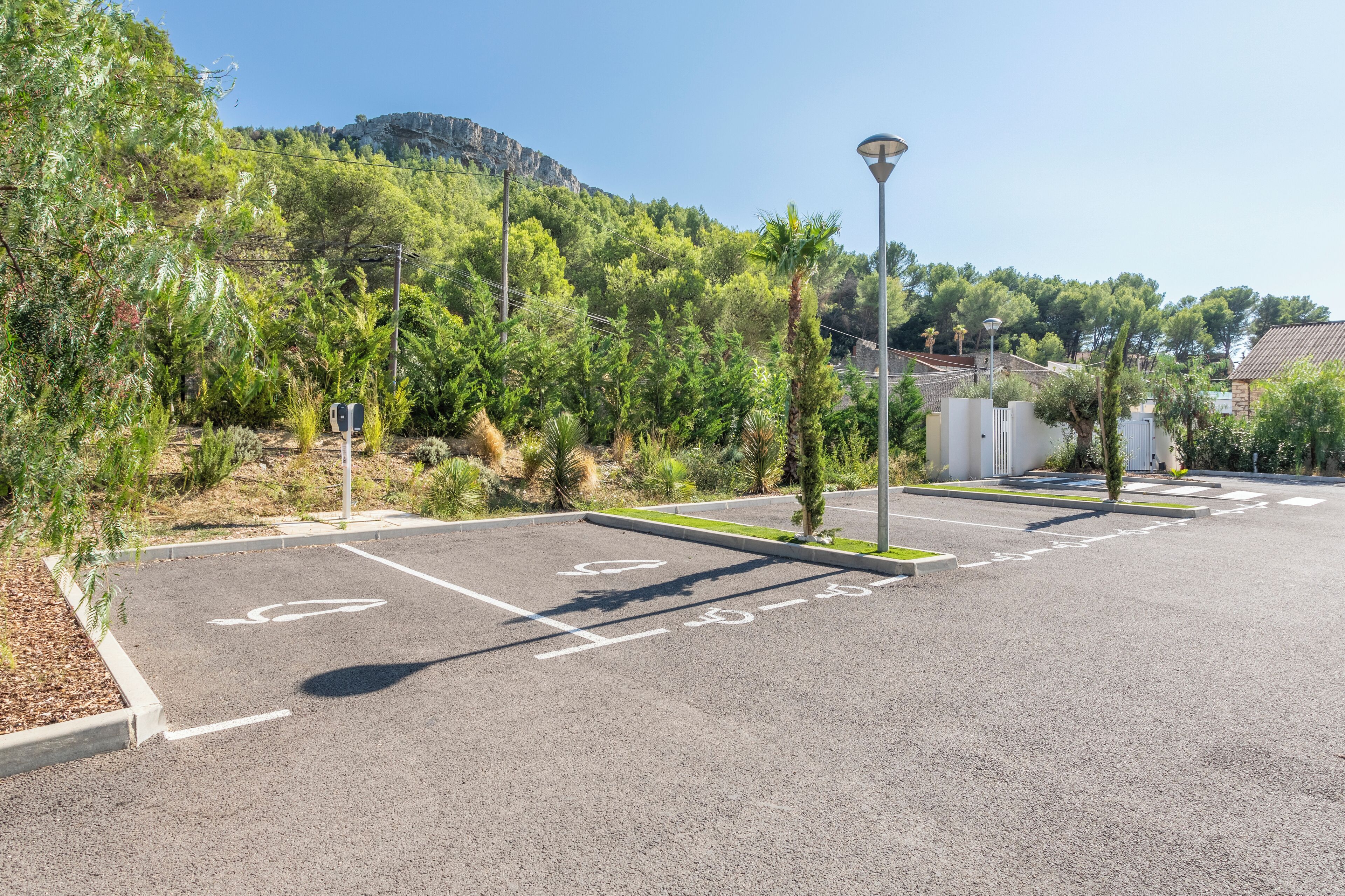 Un estacionamiento con espacios delimitados para coches y bicicletas, rodeado de vegetación y una montaña al fondo.