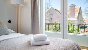 Une chambre à coucher comprenant un lit, une lampe de chevet, une fenêtre donnant sur des immeubles et un balcon avec une balustrade.