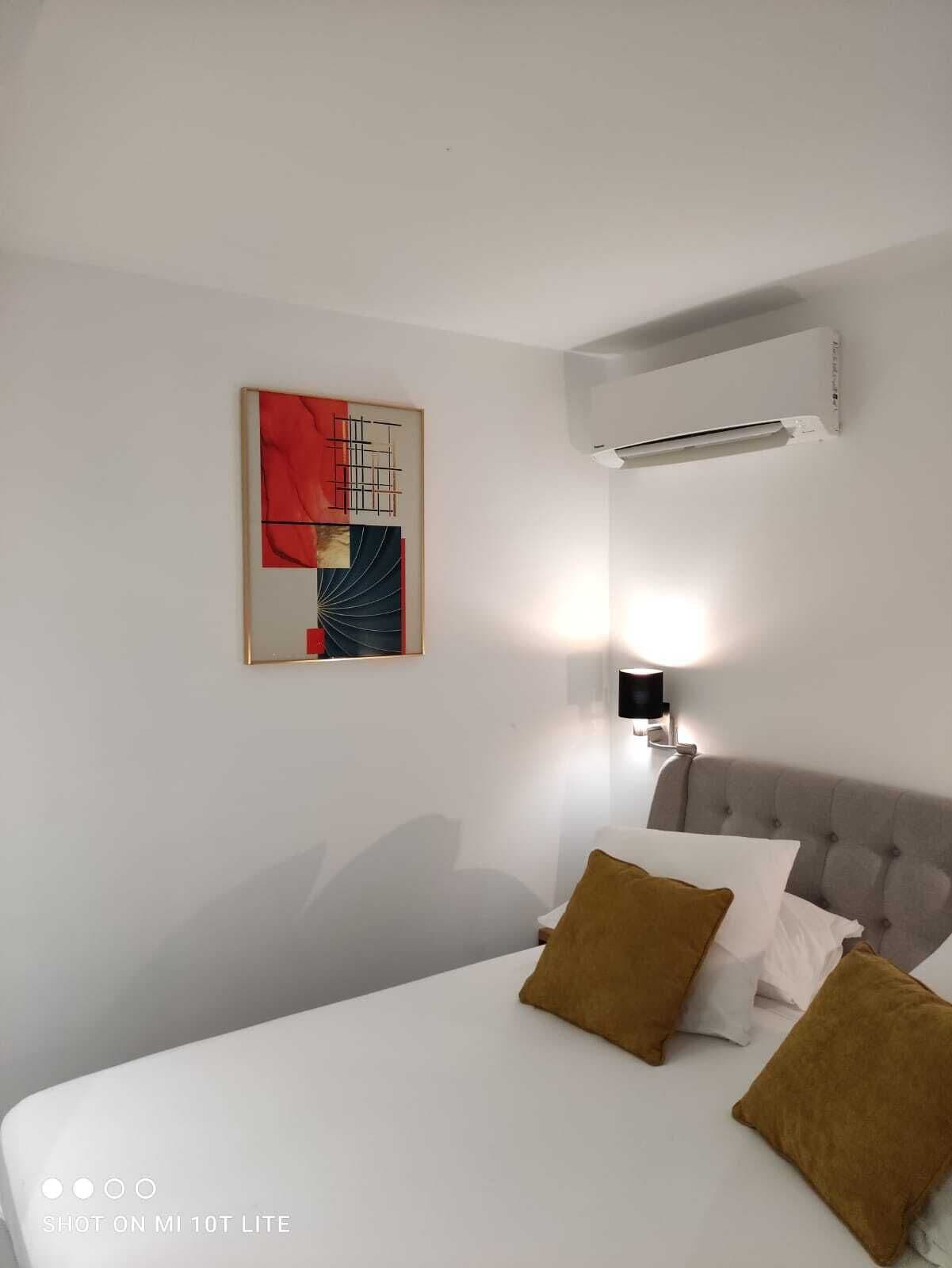 Une chambre d’hôtel avec un lit, un climatiseur fixé au mur, un tableau abstrait moderne et une lampe fixée au mur.